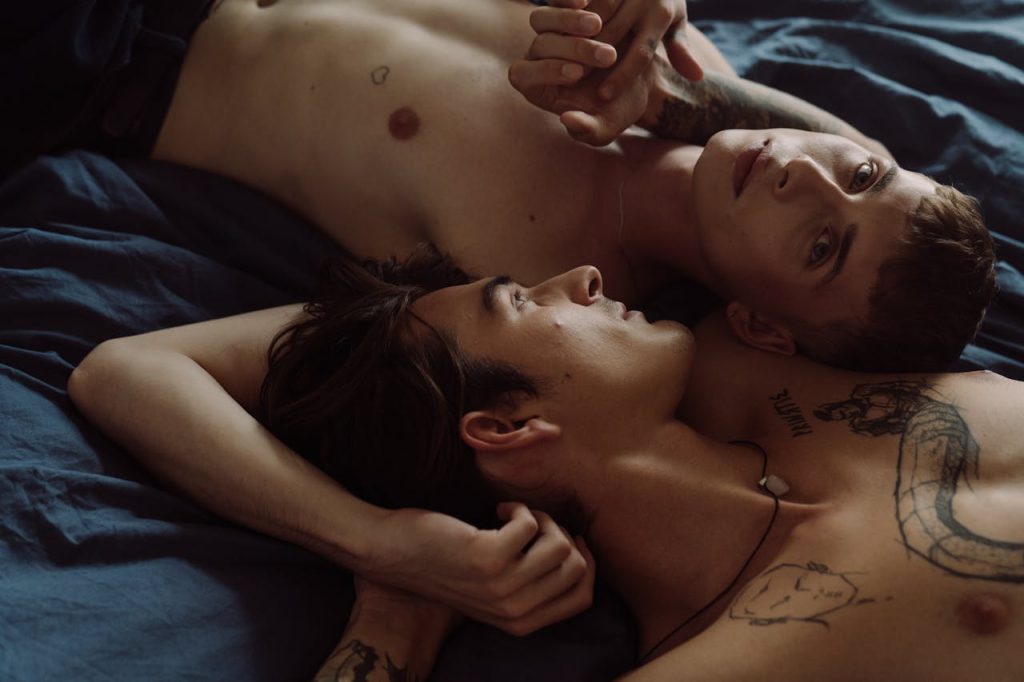 a-couple-lying-on-bed-5928595 Two tattooed men embracing intimately in a bedroom setting.